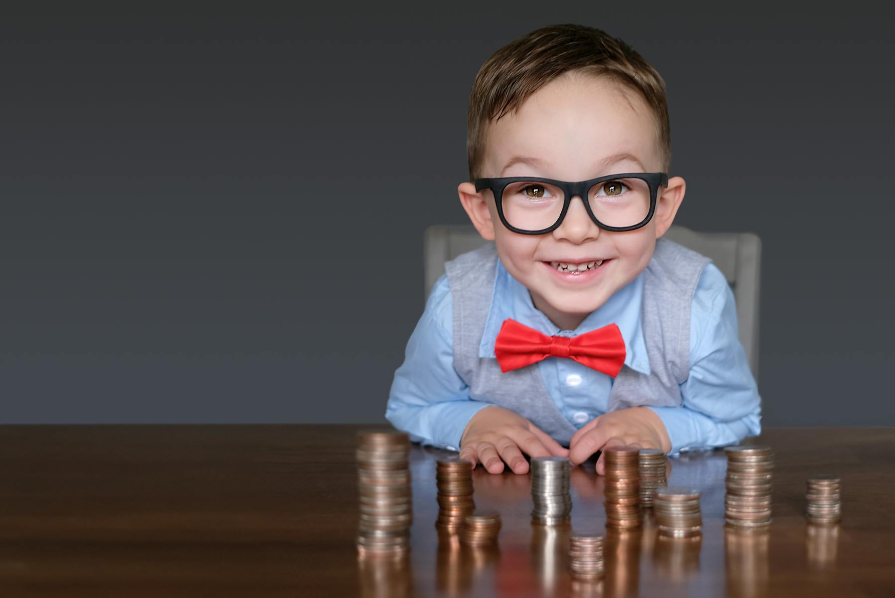 Cute kid in a bow tie and glasses smiling behind a desk with stacks of coins