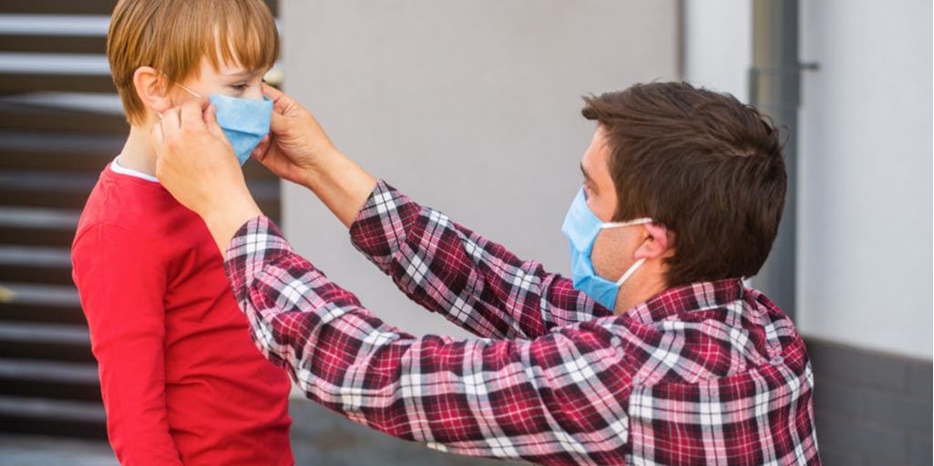 Man wearing a blue face mask and checkered shirt placing a mask on boy in red jumper